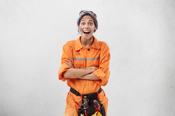 Repairing, remodeling and renovation concept. Astonished bug-eyed young female worker of maintenance service wearing orange uniform and belt kit with instruments, excited with some positive news