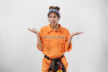 Studio portrait of attractive young European woman electrician in orange uniform, shrugging her shoulders having clueless indifferent look, saying: So what? It's not my problem. Body language
