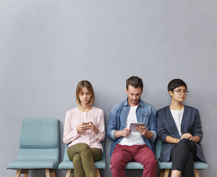 People, Employment, Recruitment, Technology Concept. Three Unemployed People Sit In Queue, Wait For Job Interview, Use Modern Gadgets, Prepare Answers On Tricky Questions From Boss Or Employer