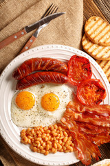 English Breakfast with sausages, grilled tomatoes, egg, bacon, beans and bread on white plate