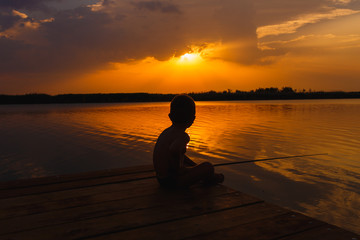 Little boy siting on wooden dock and fishing at sunset.