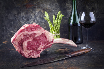 Raw dry aged wagyu tomahawk steak with green asparagus and red wine as close-up on old metal sheet