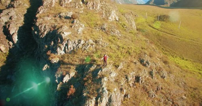 Aerial UHD 4K view. Flight from (flight to) young hiking man standing on top of the cliff at mountain meadow and rural road at sunny summer day
