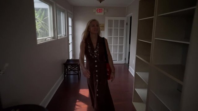 Caucasian mature woman walks out of a massage area in beautiful brown dress and goes to front desk of spa, gesticulating and laughing and shaking hands.  Steadicam shot.