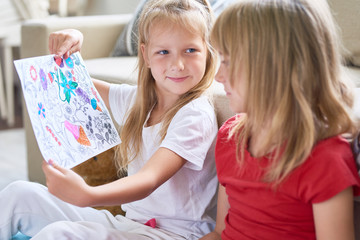 Satisfied little girl showing half-painted page from coloring book to her elder sister and waiting for her reaction, interior of cozy living room on background