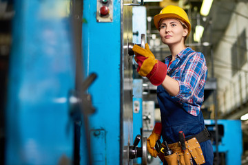 Pretty young machine operator wearing checked shirt and overall carrying out start-up works, interior of manufacturing plant on background