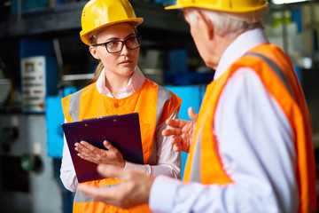 Pretty young factory worker and her senior colleague discussing results of accomplished work while standing at production department, both of them wearing hardhats and reflective jackets