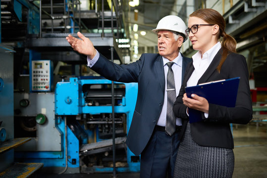 Senior entrepreneur wearing hardhat and his attractive young assistant manager carrying out inspection at production department of modern plant