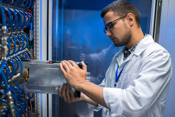 Side view portrait of young man in lab coat taking out blade server out of cabinet while working...