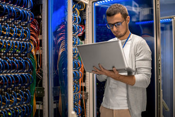Portrait of modern young man holding laptop standing in server room working with supercomputer in...