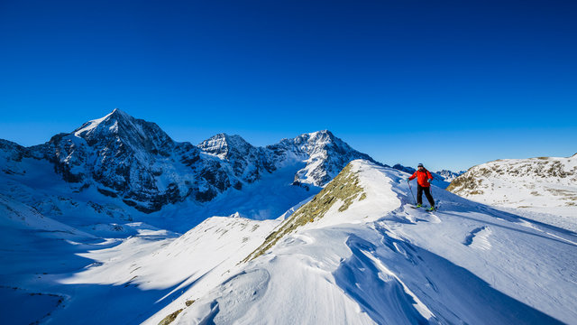 Mountain ski walking up along a snowy ridge with skis in the backpack. In background blue cloudy sky and shiny sun and Tre Cime, Drei Zinnen in South Tirol, Italy. Adventure winter extreme sport.