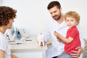 Portrait of adorable little boy looking at camera while sitting on laps of handsome bearded dentist and learning to brush teeth correctly