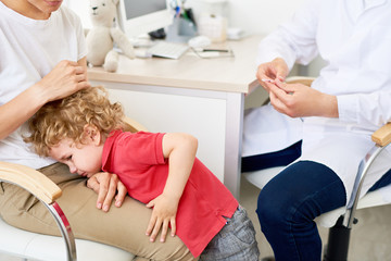 Fototapeta premium Curly little boy clinging to his mother and crying at pediatrician office refusing to be vaccinated