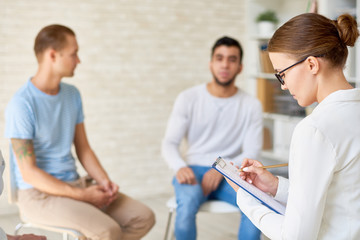 Fototapeta premium Profile view of highly professional psychiatrist taking notes on clipboard while guiding support group of young members, interior of cozy office on background
