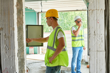 Profile view of handsome young electrician wearing hardhat and reflective jacket checking breaker box of unfinished building while his colleague talking via walkie-talkie