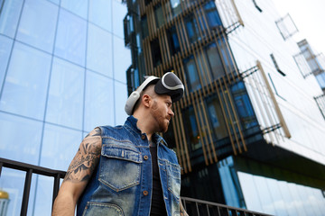 People, science, gaming, simulation and augmented reality. Serious confident young man with stubble and stylish tattoos posing in urban surroundings, using oculus rift headset for entertaining himself