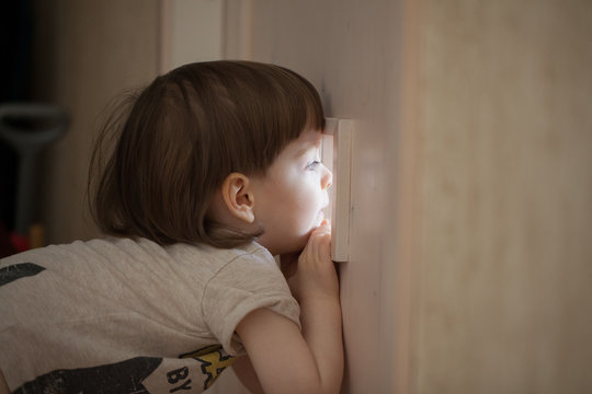 The Little Boy Looks In The Window Of The Door. A Child Spying On His Parents From Behind The Closed Bedroom Door