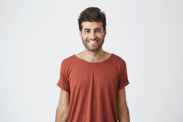 Young cheerful hispanic guy in red t-shirt brightly smiling hearing good news from friend. Beardy handsome student looking at camera with joyful smile