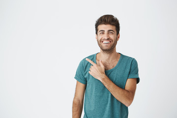 Isolated studio portrait of happy attractive caucasian guy in casual blue t-shirt, with trendy hairdo, smiling and pointing at blank wall. Copy space.
