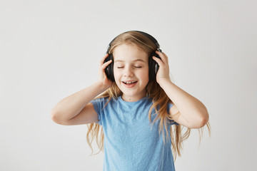 Portrait of little blonde girl in blue shirt playing with big wireless headphones, listening to music, singing song and dancing with closed eyes while nobody home.