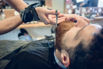 Close-up of the hand of a skilled barber using scissors, while trimming the beard of a young...