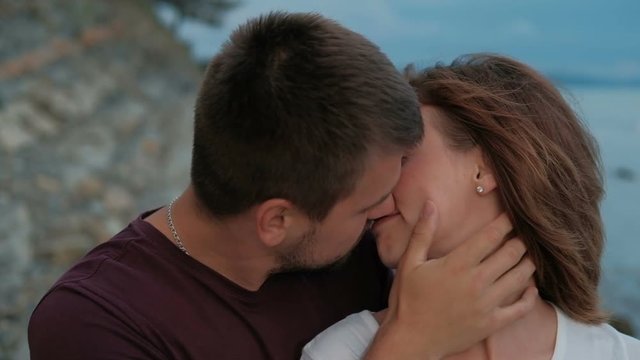 Young man kisses woman sitting on sea shore in summer evening outdoors.
