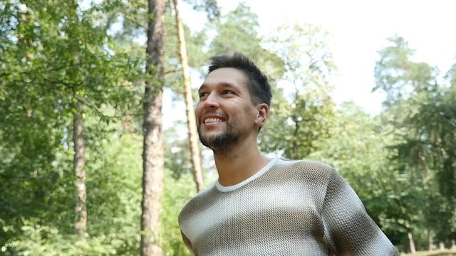 A sportive young bearded man smiles sincerely in a pine forest while standing in profile among high trees on a sunny day in summer in slow motion