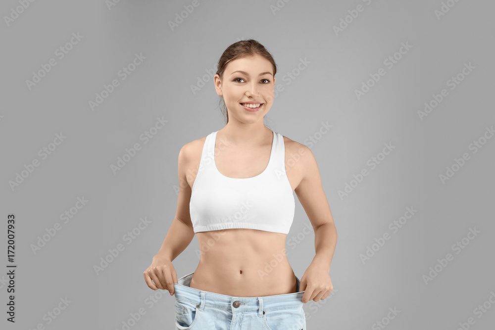 Wall mural Beautiful young woman in oversized jeans on gray background. Diet concept