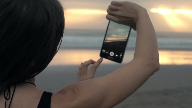 Young woman taking photo of sunset with cellphone standing on beach
