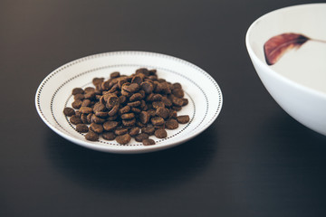White ceramic plate with kibble dry cat food placed on dark wooden floor. Close-up photo,  top view, selective focus. 