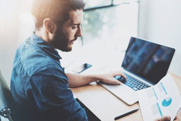 Young bearded man working at sunny loft office on laptop.Businessman analyze paper documents reports in his hand.Blurred background,horizontal.