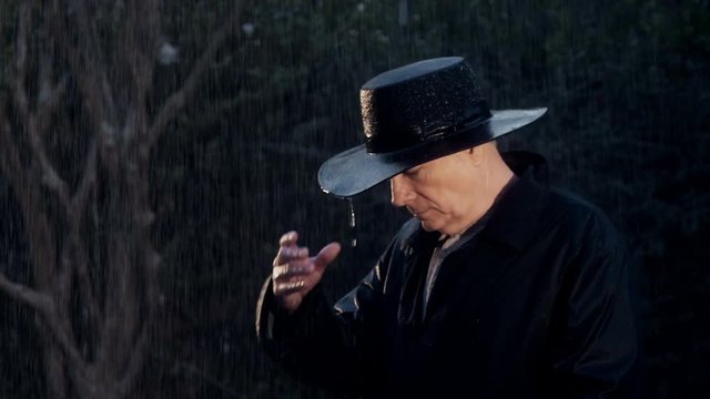 A serious mature old west type cowboy stands drenched by the rain protected only by his coat and hat. Slow motion