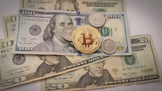 Golden physical bitcoin over US dollars and coins.  Dropping dollars and coins onto white surface.
