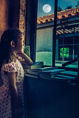Asian child standing near window and looking aside while feeling sad.
