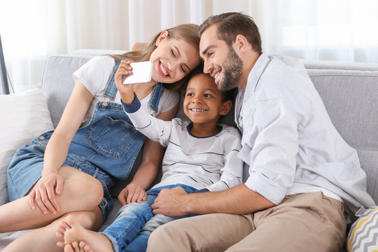 Happy couple with adopted African-American boy taking selfie while sitting on couch at home