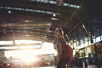 young woman in the hall of the airport terminal with small backpack as a hand luggage. silhouette of a woman in a bracing light. woman waiting for her flight on background blur airport check-In