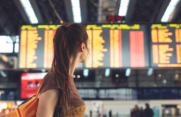 Young woman with small backpack as a hand luggage in international airport looking at the flight information board, checking her flight