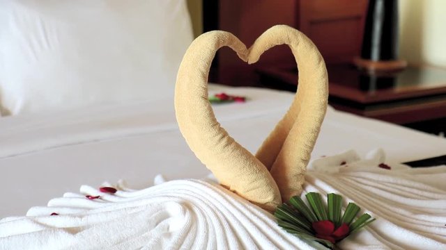 Towel as birds decoration with red rose petals on white clean bed in hotel room. Romantic bedroom for the loved couple in the hotel.