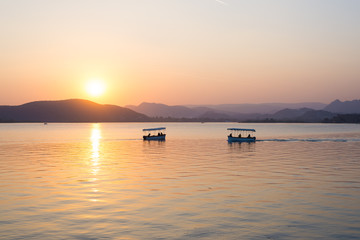 Boats floating on Lake Pichola with colorful sunset reflated on water beyong the hills. Udaipur, Rajasthan, India.