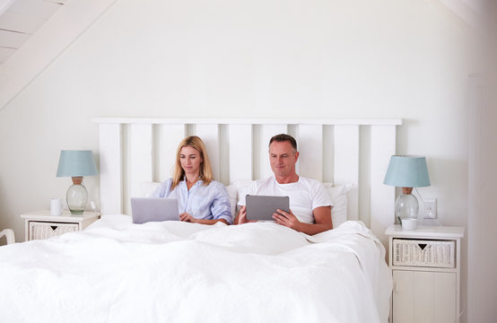 Couple Wearing Pajamas Lying In Bed Using Digital Devices