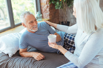 Positive aged woman giving cup of coffee to her loving husband