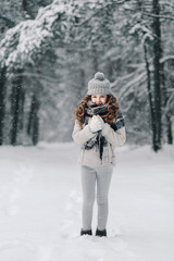 Beautiful little girl in a gray jacket with a knitted hat and scarf, walks through the park on a frosty winter day. Children play outdoors in a snow-covered forest.