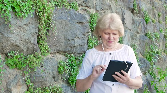 Elderly woman using tablet computer with earphones. Vintage wall of wild stone in the background.