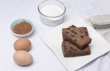 Chocolate brownie slices on a white plate with eggs, flour and coco powder
