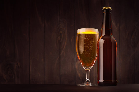 Brown beer bottle and glass tulip with golden lager on dark wood board, copy space, mock up. Template for advertising, design, branding identity.