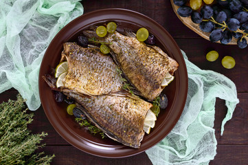 Baked carp with grapes and herbs on a ceramic dish on a dark wooden background. Top view.