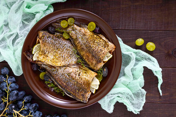 Baked carp with grapes and herbs on a ceramic dish on a dark wooden background. Top view.