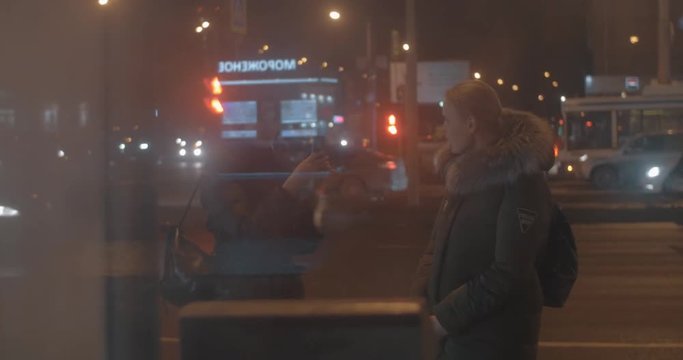 Woman taking mobile photos of the friend while they waiting for the transport in night street, view through the glass of bus stop