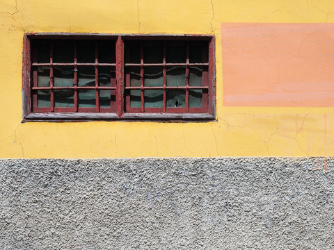 old painted yellow and grey concrete wall with narrow red framed window with bars and distressed cracked texture