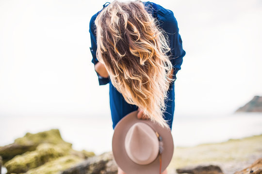 Mid adult woman in coastal setting, holding hat, bending over, hair covering face